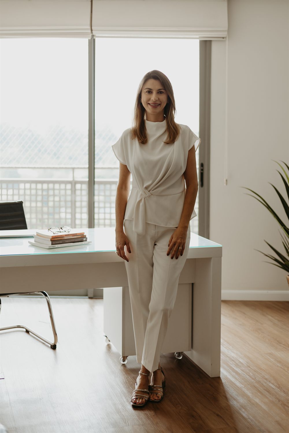 Rafaela Gualdi em pé no consultório, junto à mesa de atendimento, com luz natural ao fundo.