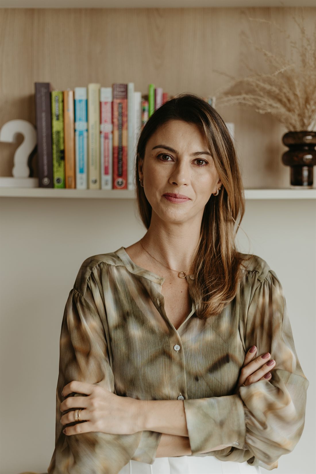 Rafaela Gualdi de braços cruzados no consultório, com uma estante de livros ao fundo.
